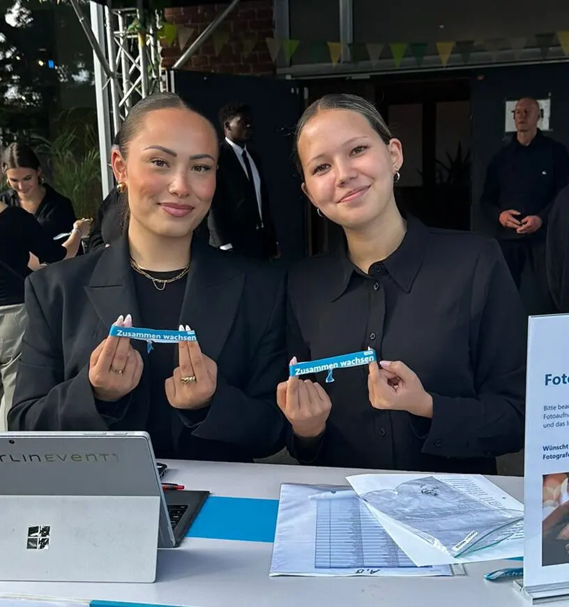 Two hostesses at a check-in desk hold up entry wristbands, with a laptop and printed lists in front of them