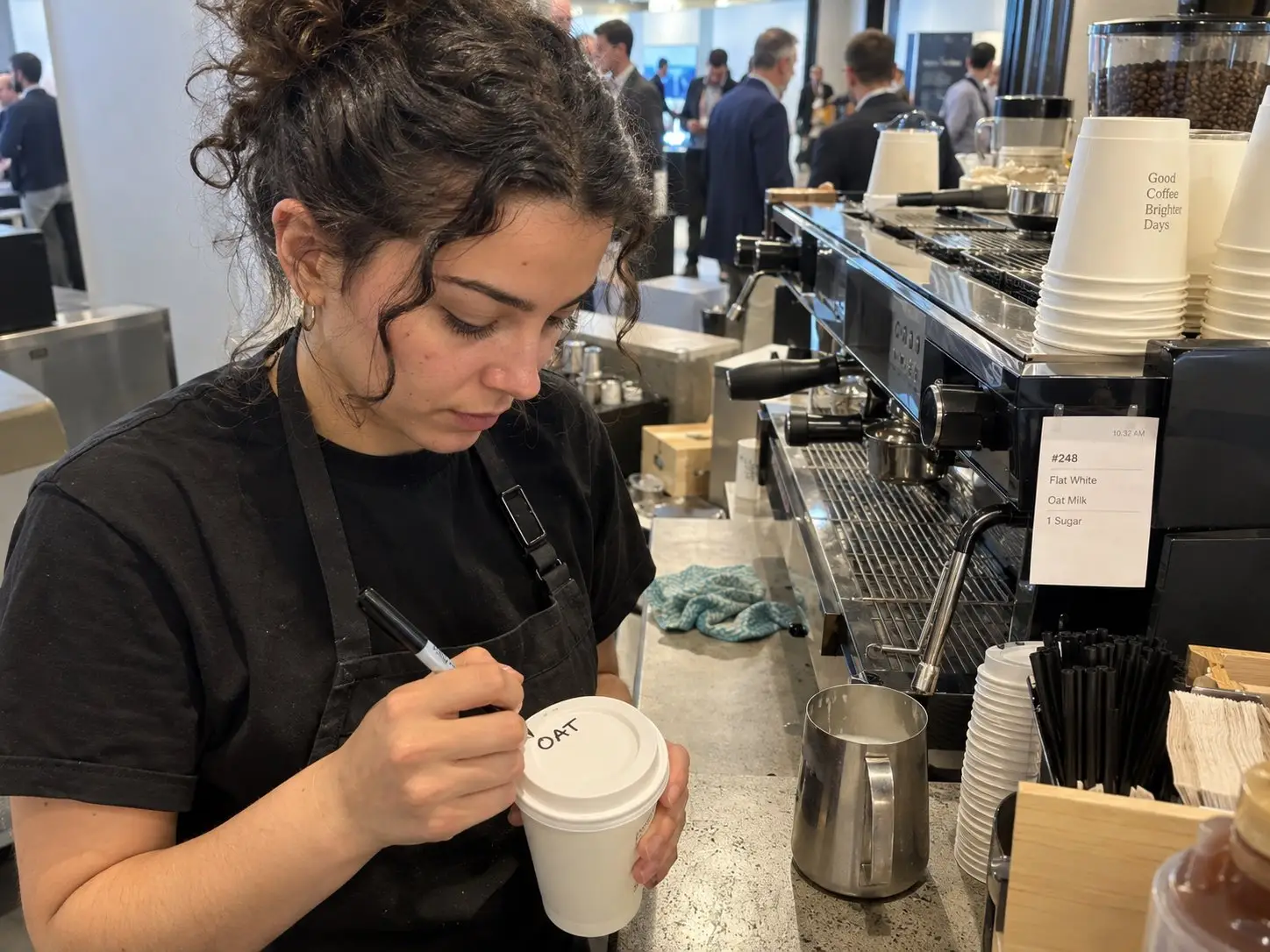 Barista beschriftet an der Kaffeebar einer Konferenz einen Becher mit „OAT“, daneben der Bon: Flat White mit Hafermilch – Barista mieten in Berlin