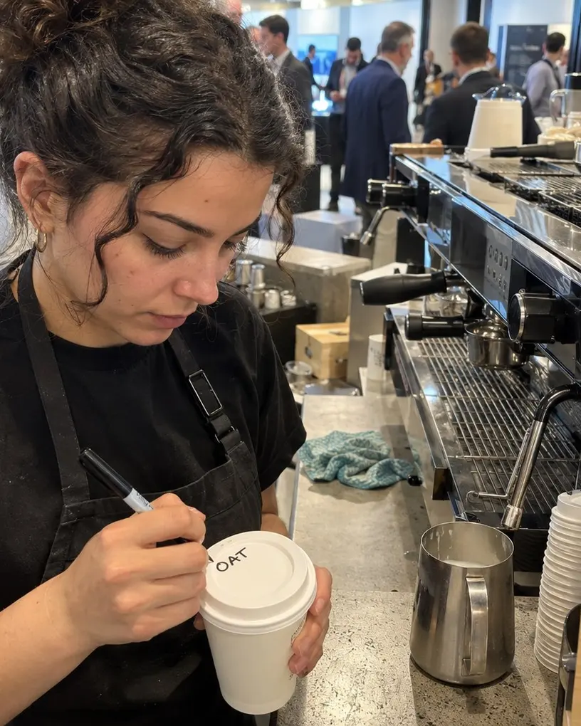 Barista marks a takeaway cup “OAT” at a coffee bar while visitors walk past behind her
