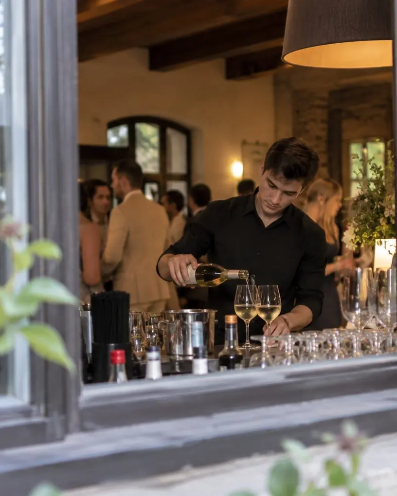 Barkeeper in Schwarz schenkt an der Fensterbar einer Gutshaus-Location Weißwein ein, drinnen feiern die Gäste – Barkeeper mieten in Berlin