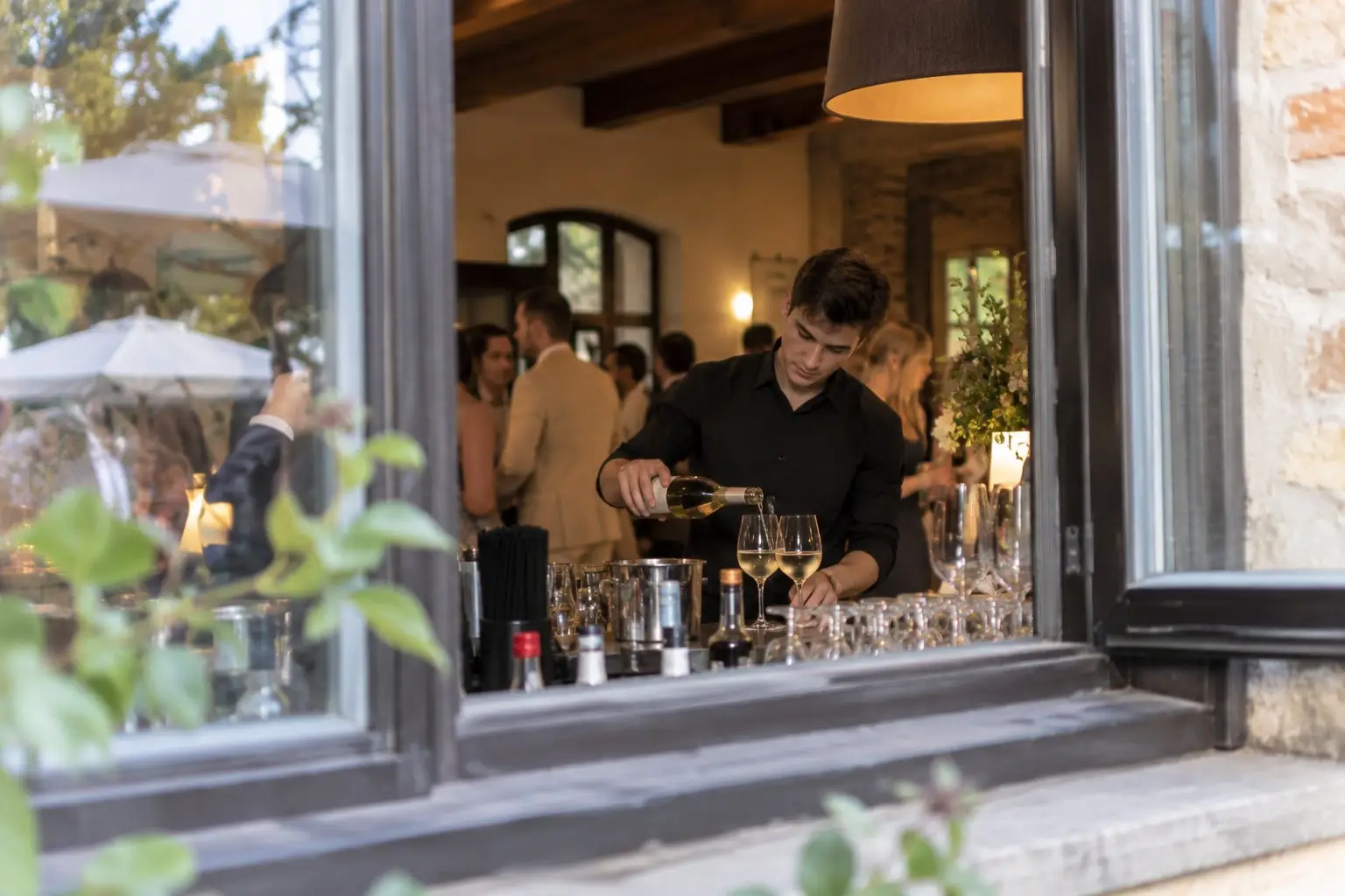 Barkeeper in Schwarz schenkt an der Fensterbar einer Gutshaus-Location Weißwein ein, drinnen feiern die Gäste – Servicepersonal für die Hochzeit