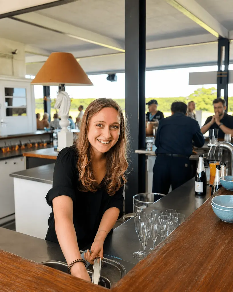 Bar staff smiles while rinsing glasses at an outdoor bar on a sunny day
