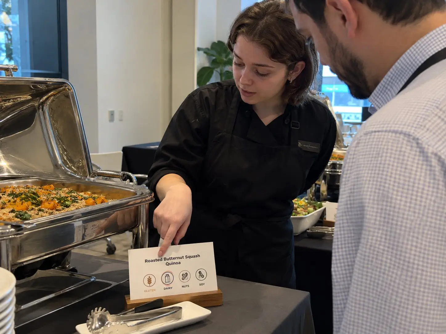 Servicekraft am Buffet erklärt einem Gast anhand des Schilds die Allergene eines Gerichts – kurzfristiges Personal in Berlin
