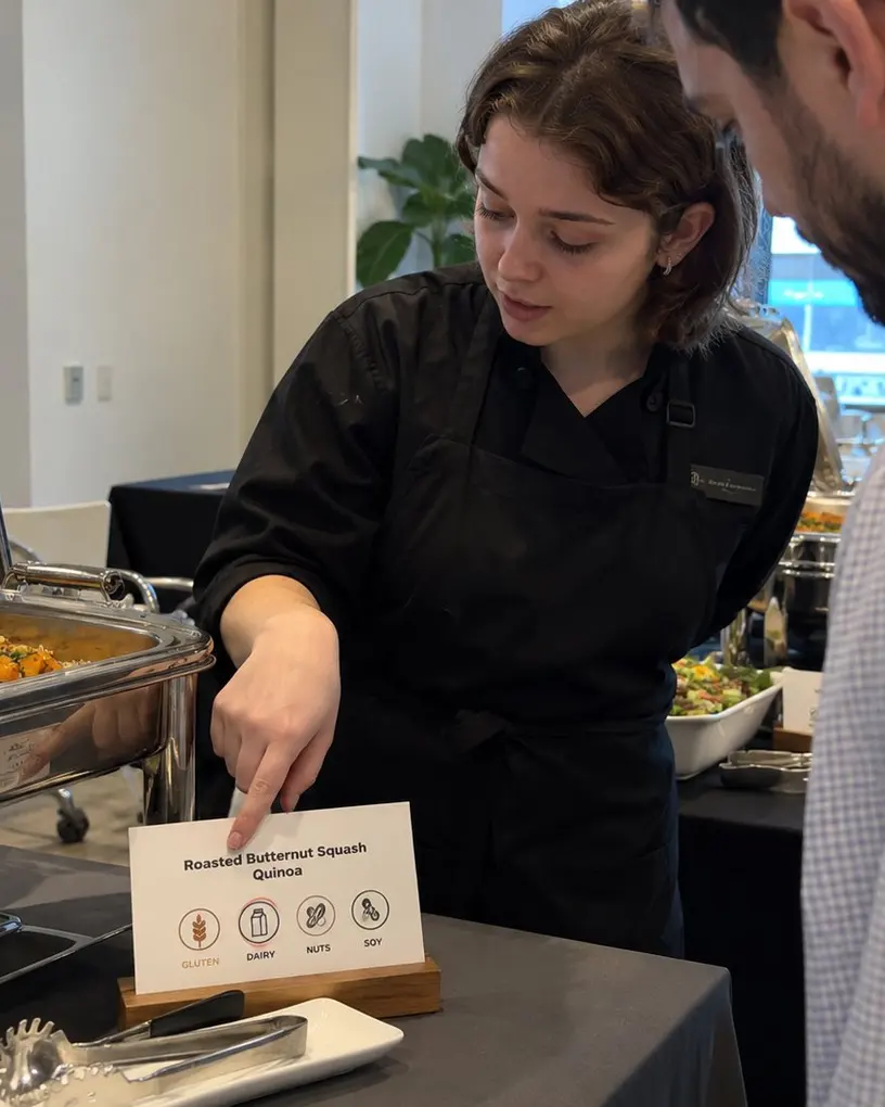Servicekraft am Buffet erklärt einem Gast anhand des Schilds die Allergene eines Gerichts – Gastronomiepersonal in Berlin
