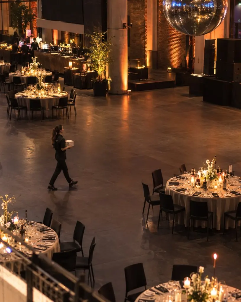Blick von der Galerie in den eingedeckten Gala-Saal mit runden Tischen, Kerzen und Bühne, eine Servicekraft geht vor dem Einlass durch den Saal – VIP-Betreuung in Berlin