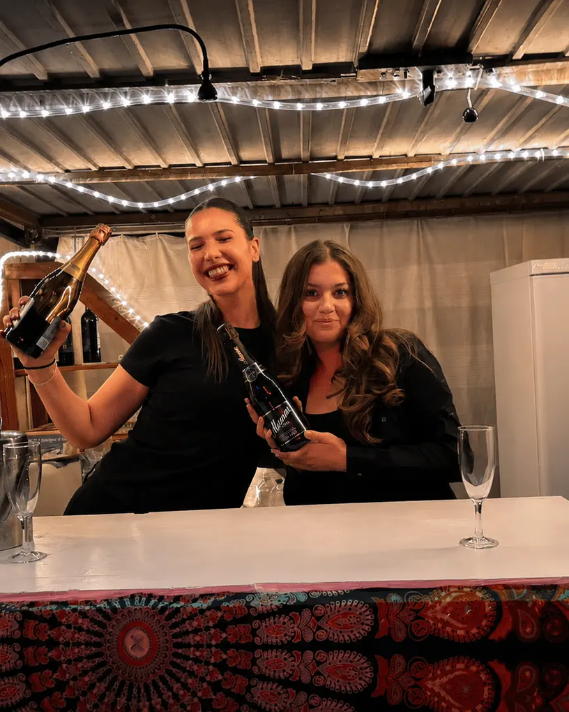 Two bar staff with bottles of sparkling wine behind a party bar hung with fairy lights