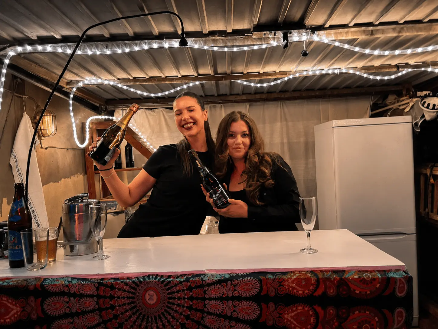 Two laughing bar staff holding bottles behind a white bar under fairy lights