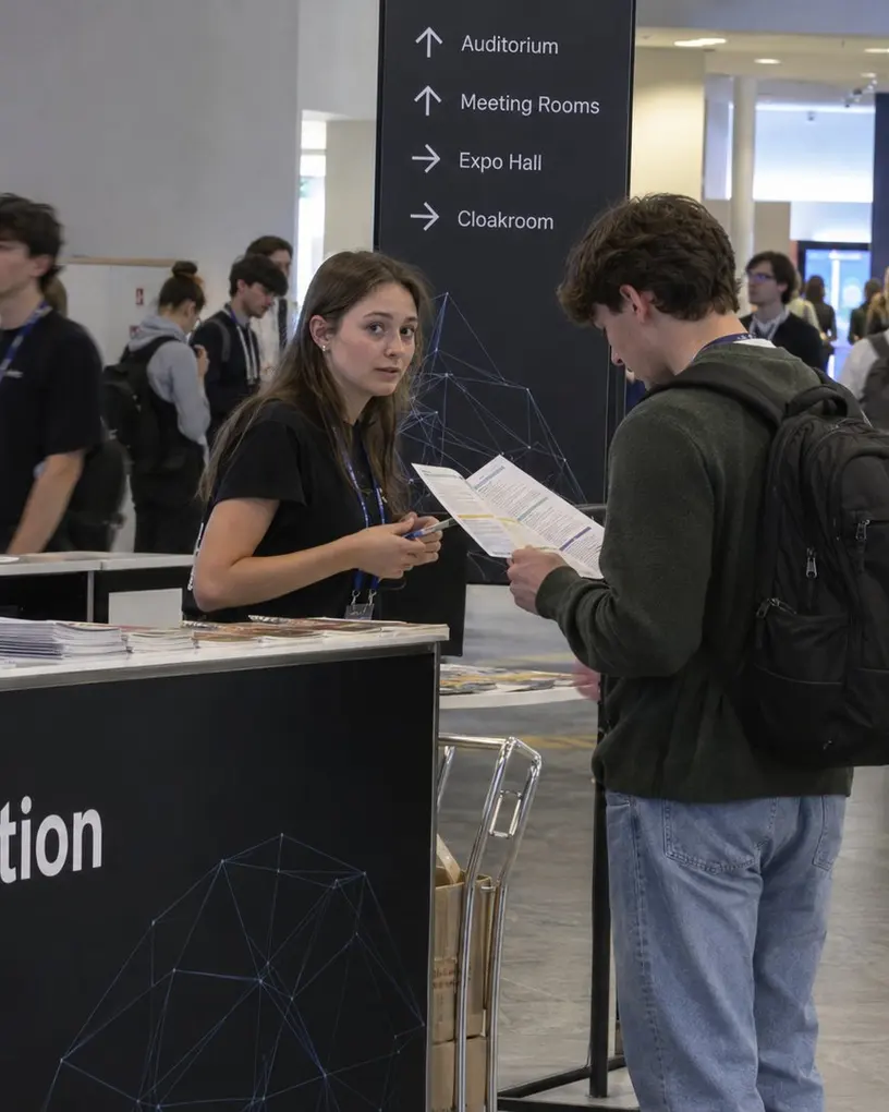 Hostess am Infocounter einer Konferenz hilft einem Teilnehmer mit dem Programmheft, dahinter die Wegweiser zu Auditorium und Expo Hall – Messepersonal für IFA, ITB und InnoTrans