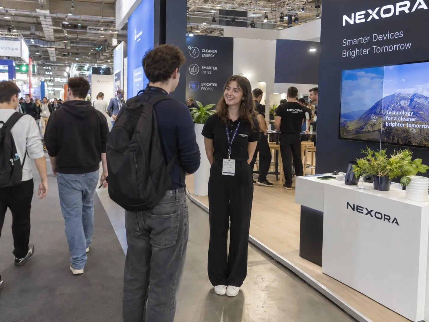 Standhostess spricht am Messestand mit einem Besucher, im Gang strömen Fachbesucher vorbei – Messepersonal in Berlin