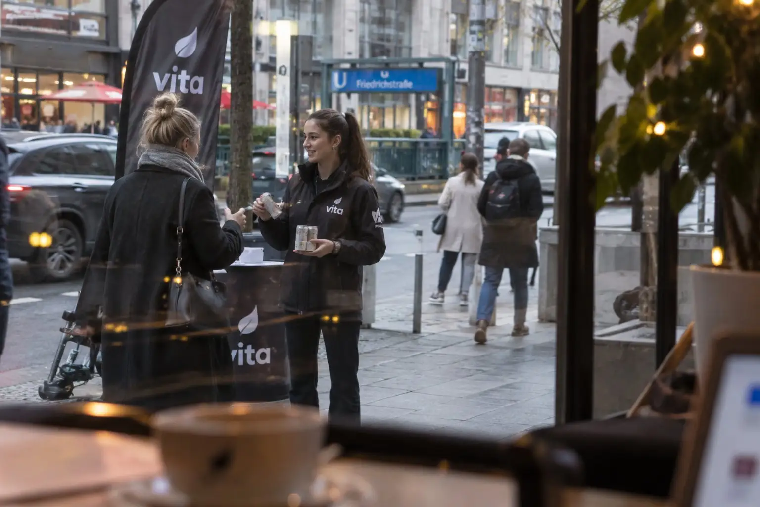 Promoterin reicht an der Friedrichstraße neben Beachflag und Stehtisch einer Passantin eine Produktprobe, gesehen aus einem Café – Sampling-Personal für Roadshows