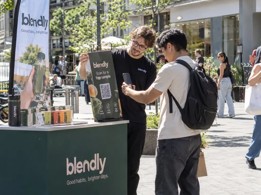Promoter zeigt Passanten an einem Sampling-Stand einen QR-Code und reicht eine Produktprobe